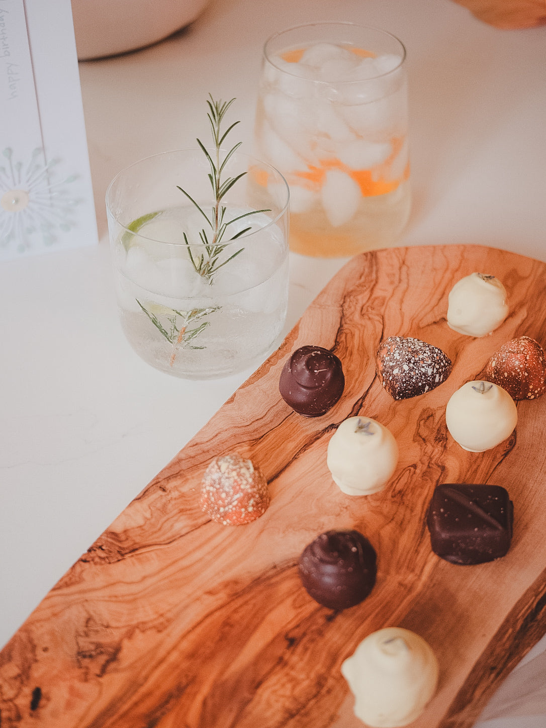 Wooden board with chocolates and a glass of rosemary cocktail on a white kitchen surface