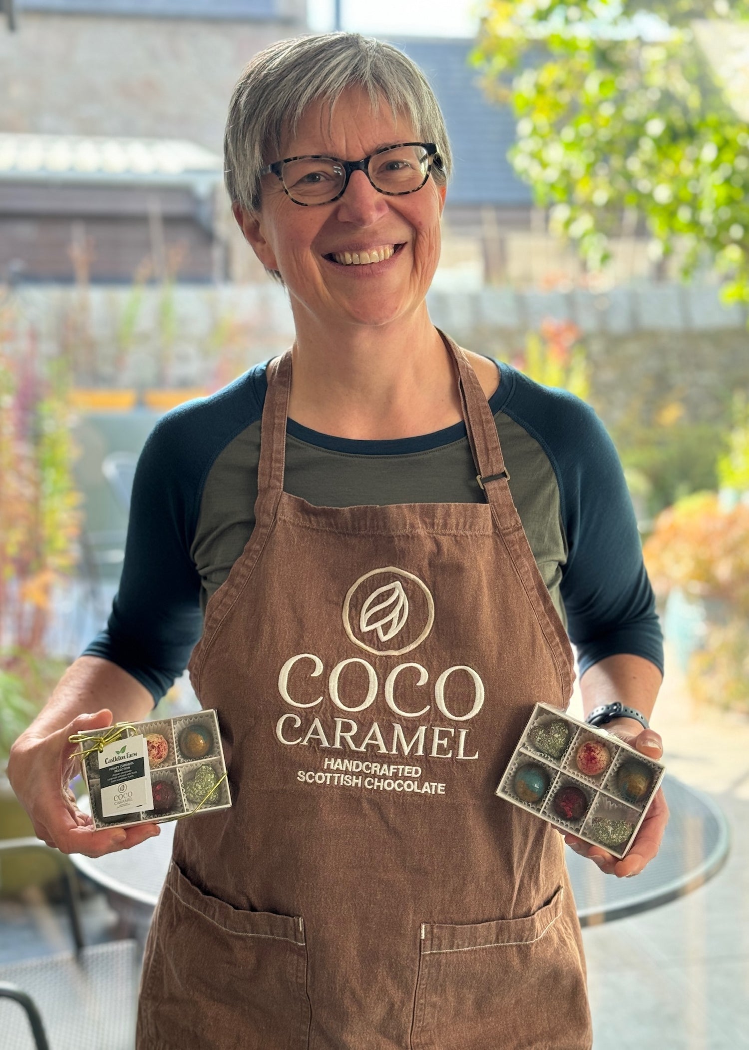 Person wearing a 'Coco Caramel' apron holding chocolate boxes outdoors.