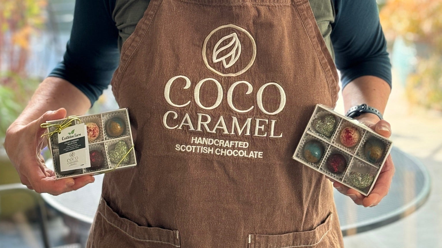 Helen wearing a 'Coco Caramel' apron holding chocolate boxes outdoors.