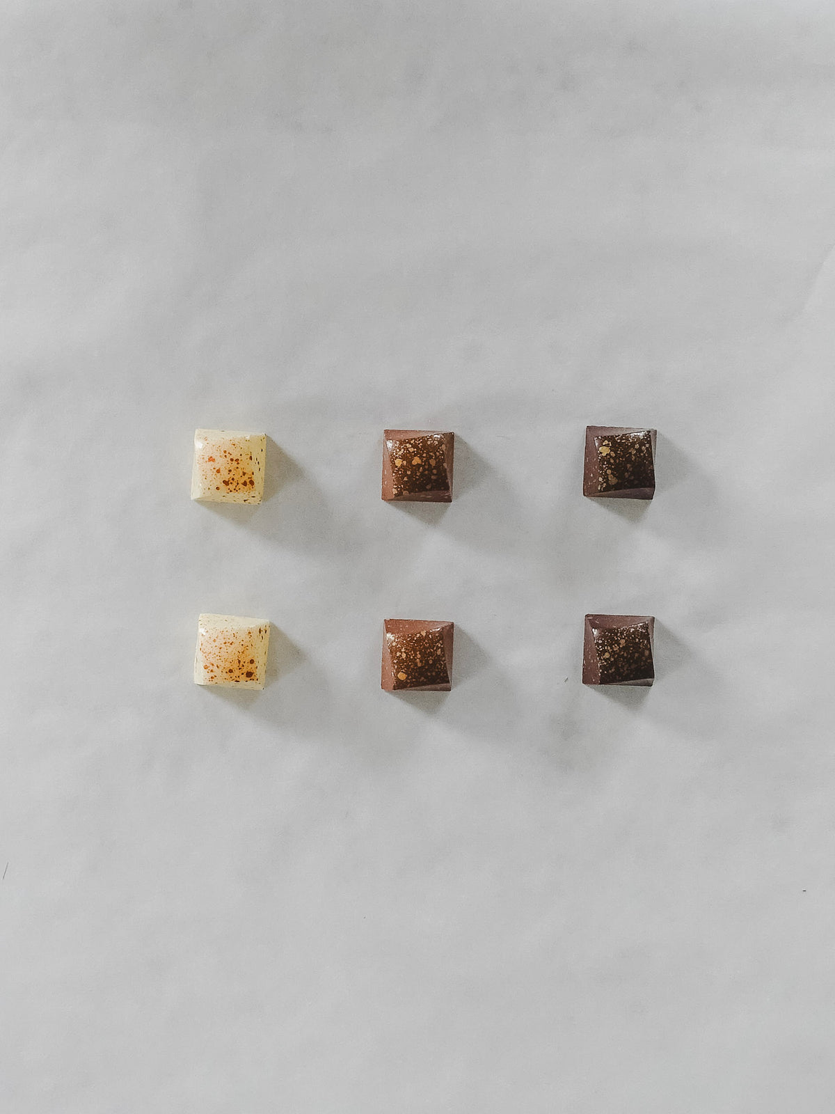 Six small square chocolate pieces arranged in two rows on a light gray marble surface. Three are milk chocolate brown and three are dark chocolate brown, with one white chocolate piece featuring orange-red sprinkles.