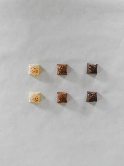 Six small square chocolate pieces arranged in two rows on a light gray marble surface. Three are milk chocolate brown and three are dark chocolate brown, with one white chocolate piece featuring orange-red sprinkles.